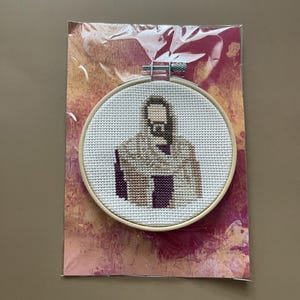 Puede incluir: Un bastidor de bordado circular de madera que contiene un patr&oacute;n de punto de cruz de una figura con barba, vistiendo una t&uacute;nica color canela sobre una prenda de color morado oscuro. El bordado est&aacute; sobre tela blanca y en una funda pl&aacute;stica.