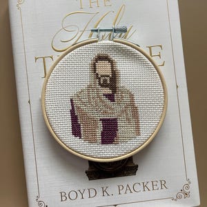 Puede incluir: Un bastidor de bordado circular de madera que contiene un dise&ntilde;o de punto de cruz de una figura con cabello largo casta&ntilde;o y barba, vistiendo una t&uacute;nica color canela y morado oscuro. La tela es Aida blanca y tiene un tornillo de metal arriba.