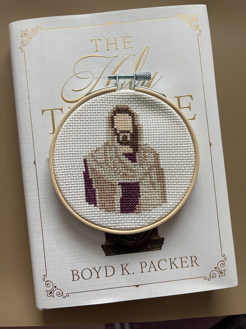 Puede incluir: Un bastidor de bordado circular de madera que contiene un dise&ntilde;o de punto de cruz de una figura con cabello largo casta&ntilde;o y barba, vistiendo una t&uacute;nica color canela y morado oscuro. La tela es Aida blanca y tiene un tornillo de metal arriba.