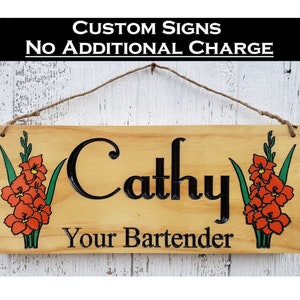 May include: A wooden sign with the name "Cathy" and the text "Your Bartender" in black lettering. The sign is decorated with two orange flowers with green leaves on each side.