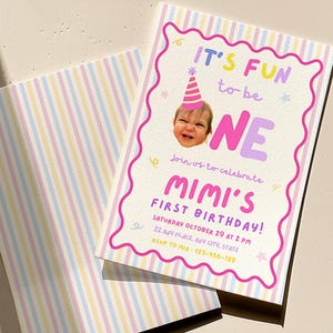 May include: A rectangular birthday invitation card with a pastel striped envelope. The card features a photo of a child in a pink party hat, the text IT'S FUN to be ONE, MIMI'S FIRST BIRTHDAY, and event details in colourful, playful lettering.