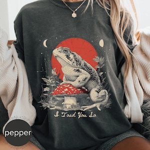 May include: Dark grey t-shirt with a detailed illustration of a toad on a red and white mushroom, set against a red sun and crescent moons. The text "I Toad You So" is below the toad. The shirt has a vintage style.