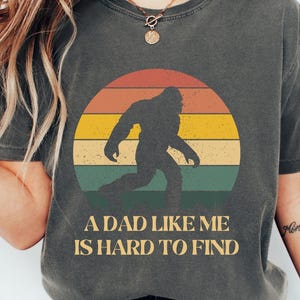 May include: Dark gray t-shirt featuring a Bigfoot silhouette in front of a retro sunset design. The text "A DAD LIKE ME IS HARD TO FIND" is printed below the graphic. The shirt is a casual wear item.
