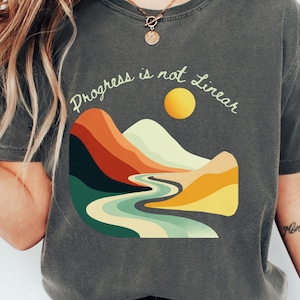 May include: Dark gray t-shirt with the text "Progress is not Linear" above a colorful landscape illustration. The graphic features mountains, a river, and a yellow sun. The shirt is a crew neck style.