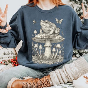May include: Navy blue sweatshirt featuring a detailed illustration of a toad sitting on a mushroom, with butterflies and the text "let me be weird in peace." The design is in a vintage style.