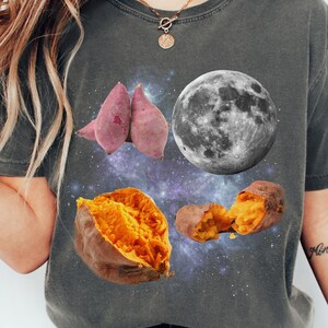 May include: Dark grey t-shirt featuring a graphic design of sweet potatoes, a full moon, and a starry galaxy background. The sweet potatoes are in various forms, including whole and cut open, with vibrant orange flesh.