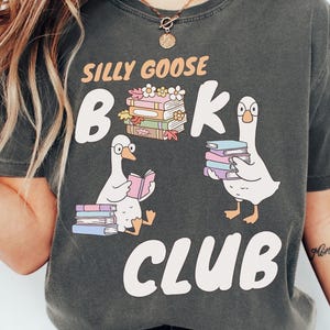 Può includere: T-shirt grigio scuro con le parole "Silly Goose Book Club" in bianco. La maglietta presenta oche dei cartoni animati e pile di libri in rosa, viola e verde. Le oche tengono e leggono libri.
