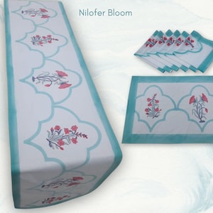 Può includere: Un set di runner da tavolo, tovaglietta e tovaglioli bianchi con un motivo floreale. Il set presenta un bordo azzurro e un motivo floreale in rosso e viola. Il testo "Nilofar Bloom" si trova nell'angolo in alto a destra.
