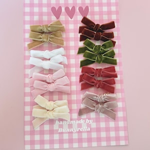 May include: A collection of velvet hair bows in various colors, including beige, brown, white, pink, and green, arranged on a pink and white checkered background. The bows are handmade by Bunnyrella.