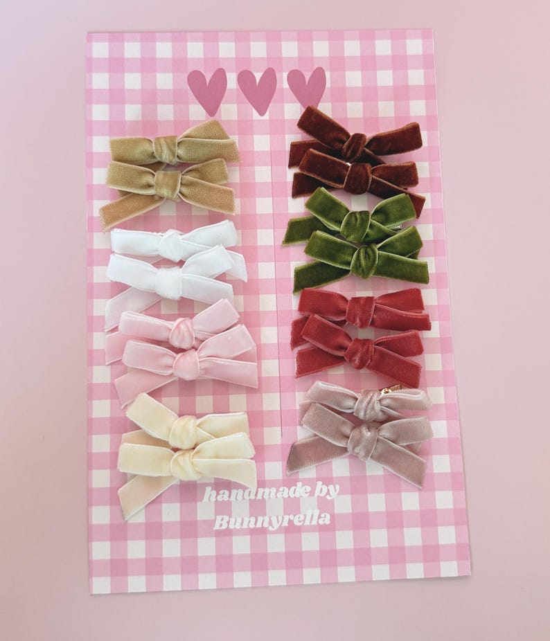 May include: A collection of velvet hair bows in various colors, including beige, brown, white, pink, and green, arranged on a pink and white checkered background. The bows are handmade by Bunnyrella.
