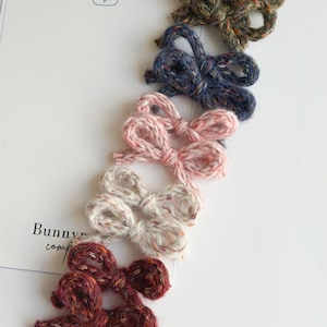 May include: Five handmade yarn bows in various colors: olive green, navy blue, light pink, off-white, and burgundy. The bows are arranged on a white surface with a bunny illustration and the text "Bunny comp".