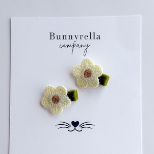 May include: Two handmade flower hair clips. Each clip features a cream-colored, crocheted flower with a brown center, attached to a green barrette. The clips are displayed on a white card with the text "Bunnyrella company" and a cat face illustration.