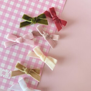 May include: An assortment of velvet hair bows in various colors, including pink, green, and white, are arranged on a pink and white checkered background. The bows are tied in a classic bow shape. Three pink hearts are visible at the top of the background.