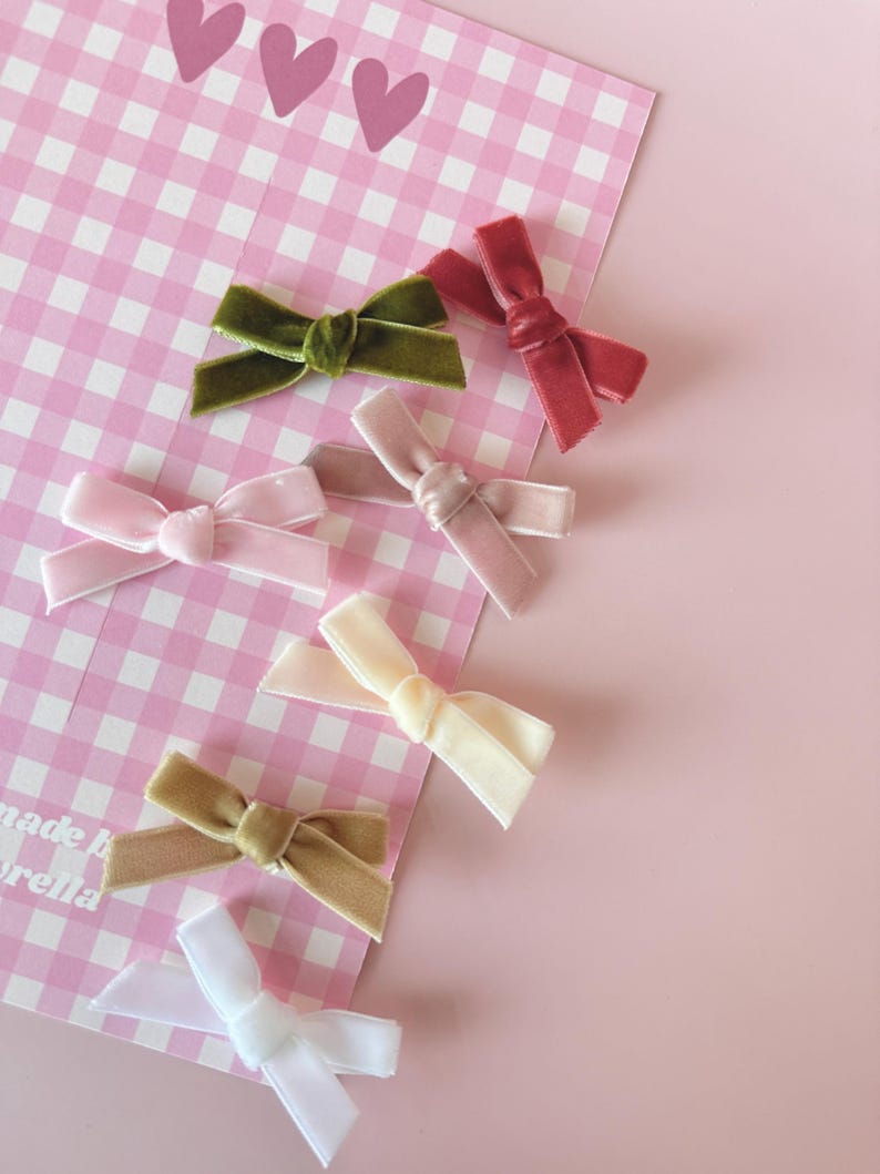 May include: An assortment of velvet hair bows in various colors, including pink, green, and white, are arranged on a pink and white checkered background. The bows are tied in a classic bow shape. Three pink hearts are visible at the top of the background.