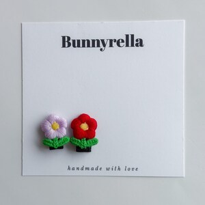 Może przedstawiać: Biała karta z napisem "Bunnyrella" i "handmade with love". Dołączone są dwie haftowane spinki do włosów w kształcie kwiatów. Jedna jest fioletowa z żółtym środkiem, a druga czerwona z żółtym środkiem. Obie mają zielone liście.