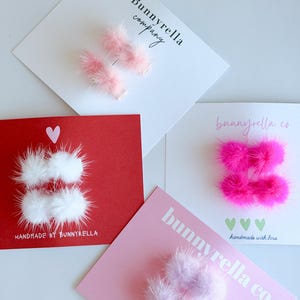 May include: A collection of handmade hair clips in various colors, including pink, white, and purple, arranged on cards. The cards feature the text "Bunnyrella company" and "handmade with love."