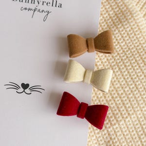 May include: Three small velvet bow hair clips in brown, cream, and red are displayed on a white card with the text "Bunnyrella company". The card also features a cat face graphic. The background is a cream-colored knitted fabric.