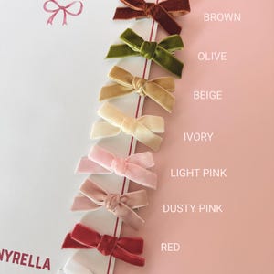 May include: A collection of velvet hair bows in various colors, including brown, olive, beige, ivory, light pink, dusty pink, red, and white. The bows are arranged on a white card with the brand name "NYRELLA" visible.