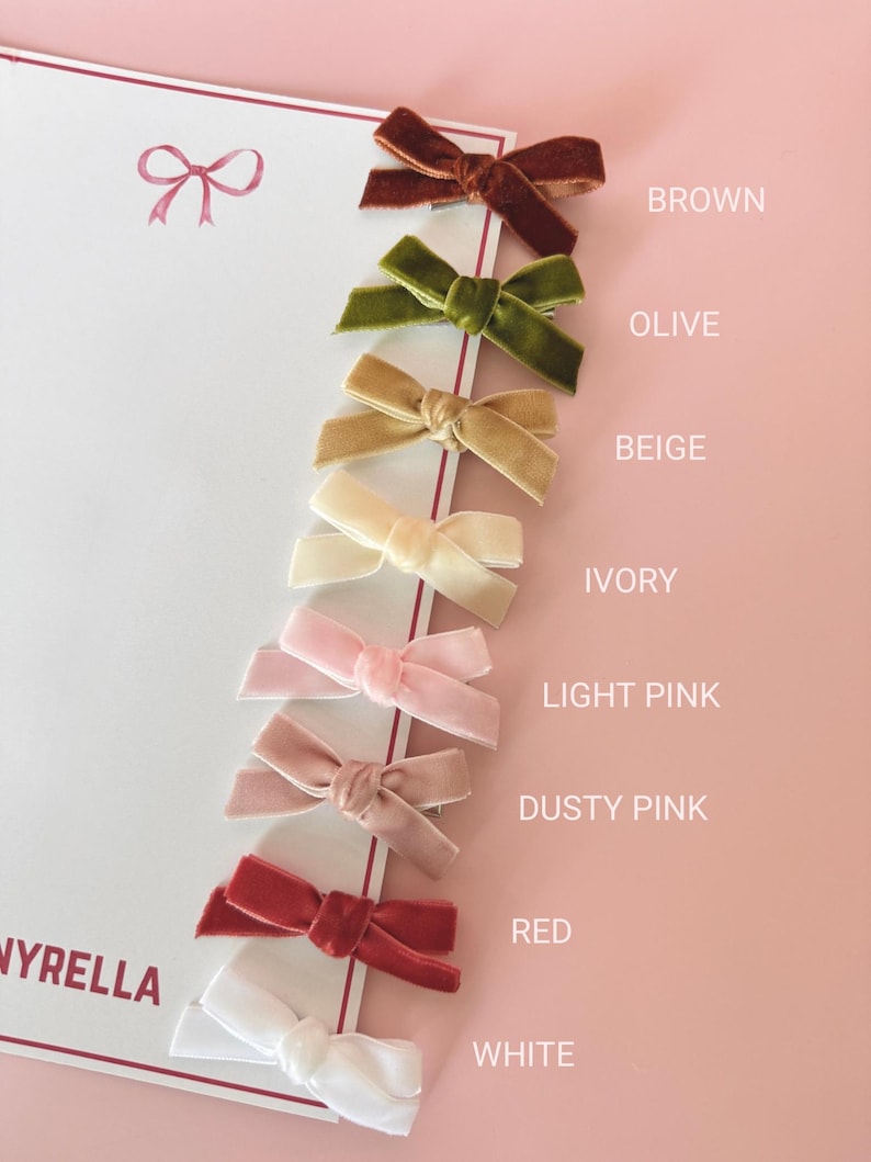 May include: A collection of velvet hair bows in various colors, including brown, olive, beige, ivory, light pink, dusty pink, red, and white. The bows are arranged on a white card with the brand name "NYRELLA" visible.