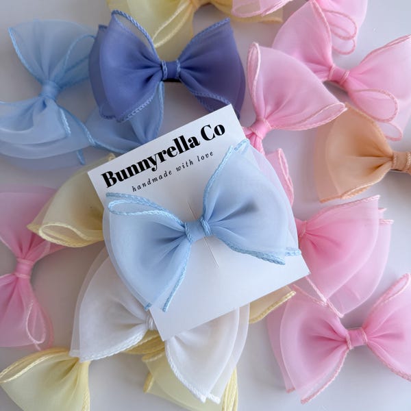 Puede incluir: Colección de lazos de tela transparente en tonos pastel, rosa, amarillo, azul y blanco. Una tarjeta con el texto "Bunnyrella Co handmade with love" está en el centro. Accesorios para el cabello.