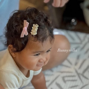 May include: A baby with dark curly hair wearing a white onesie. Two hair clips adorn the baby's hair: a light pink bow and a gold flower clip. The brand name "Bunnyrella" is visible in the image.