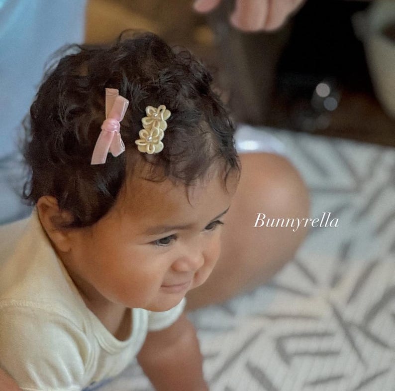 May include: A baby with dark curly hair wearing a white onesie. Two hair clips adorn the baby's hair: a light pink bow and a gold flower clip. The brand name "Bunnyrella" is visible in the image.