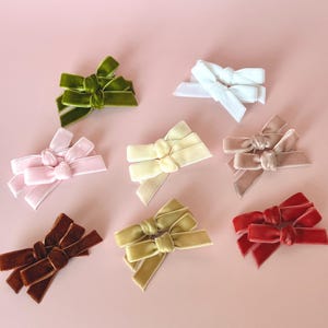 May include: A collection of velvet hair bows in various colors, including green, white, pink, yellow, taupe, brown, olive green, and red. The bows are arranged on a light pink background, showcasing their textures and hues.