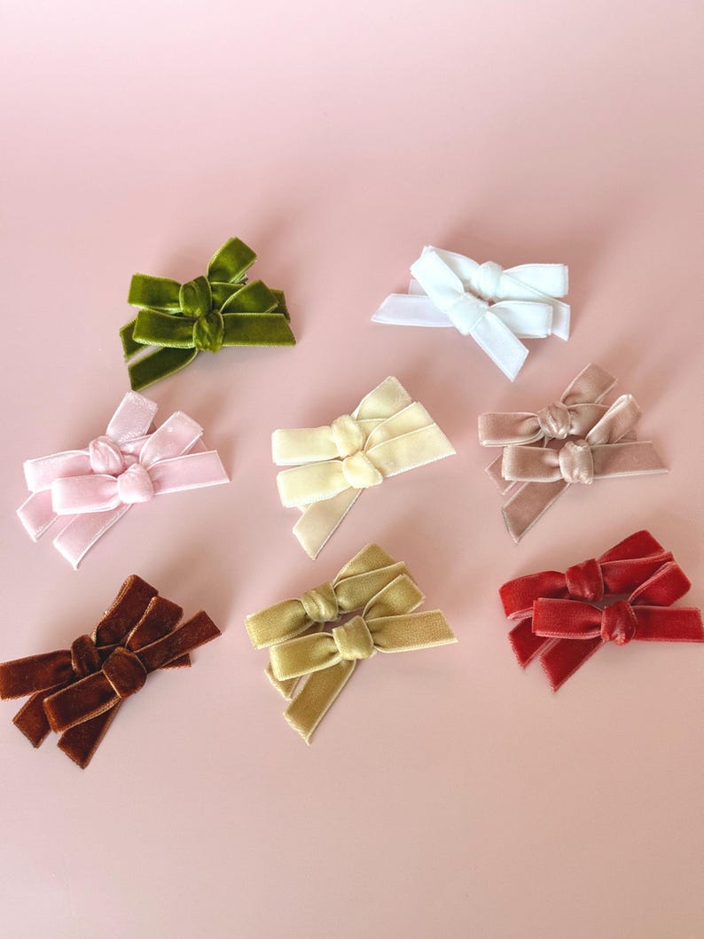 May include: A collection of velvet hair bows in various colors, including green, white, pink, yellow, taupe, brown, olive green, and red. The bows are arranged on a light pink background, showcasing their textures and hues.
