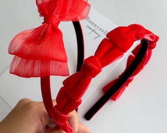May include: Two red headbands with bows. One headband has a large, layered bow made of sheer fabric. The other headband has a velvet bow. The headbands are on a white surface. The text "Bunnyrell company" is visible.