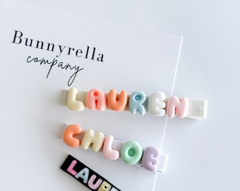 May include: Three hair clips with names. Two clips have pastel-colored letters spelling "LAUREN" and "CHLOE" on white ribbon. A third clip has "LAUREN" in pastel letters on a black background. The Bunnyrella company logo is visible.