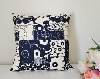May include: Square decorative pillow with a patchwork design featuring large, stylized floral patterns in navy blue and cream. The pillow is placed on a white surface, with a bouquet of flowers in the background.