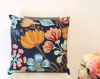 Puede incluir: Un cojín decorativo cuadrado azul marino con un diseño floral bordado. Las flores son en tonos naranja, rosa y azul, con tallos y hojas marrones. El cojín está colocado sobre una silla blanca.