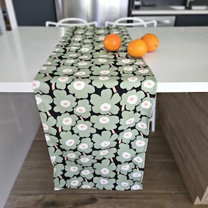 Peut inclure: Chemin de table floral vert et noir avec un motif répété de grandes fleurs. Les fleurs sont vert clair avec des centres blancs et pêche. Trois oranges reposent sur la surface blanche. Le chemin de table est en tissu.