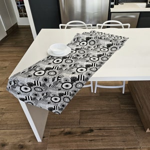 Puede incluir: Un camino de mesa con estampado floral en blanco y negro se muestra sobre una mesa blanca. El camino de mesa presenta un diseño repetido de flores estilizadas y formas geométricas. Un cuenco y un plato blancos están sobre la mesa.