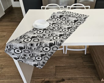Könnte beinhalten: Ein schwarz-weiß gemusterter Tischläufer liegt auf einem weißen Tisch. Der Läufer hat ein sich wiederholendes Muster aus stilisierten Blumen und geometrischen Formen. Eine weiße Schüssel und ein Teller stehen auf dem Tisch.