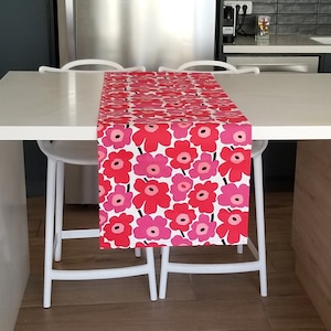 Puede incluir: Un camino de mesa floral rojo y rosa sobre una mesa blanca. El diseño presenta un patrón repetido de grandes flores estilizadas en tonos rojos y rosas sobre un fondo blanco. La mesa está rodeada de sillas blancas.
