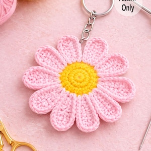Pode incluir: Um chaveiro de margarida de crochê, com centro amarelo e pétalas rosa. O chaveiro é preso a um anel e corrente de metal prateado. As palavras "Pattern Only" são visíveis em um círculo branco. O fundo é rosa claro.