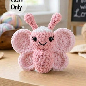 Puede incluir: Una mariposa rosa hecha a mano con ojos negros y una boca sonriente. La mariposa tiene alas grandes y redondeadas y antenas. El texto "Pattern Only" está en un círculo blanco en la esquina superior izquierda. La mariposa está sobre una superficie de madera.