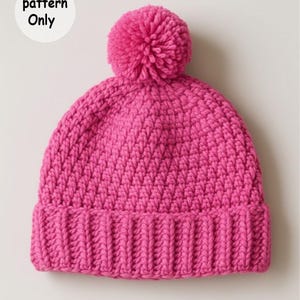 以下が含まれることがあります： 明るいピンク色のニット帽。リブ編みのつばと大きなポンポンが付いています。帽子は、テクスチャ加工された斜めのステッチパターンが特徴です。「pattern Only」というテキストが左上の白い円の中にあります。