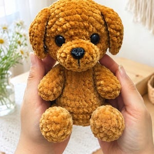 May include: A small, golden-brown plush dog toy crafted from soft, textured yarn. The stuffed animal features round, black plastic eyes and a small, black plastic nose, with floppy ears and a seated posture, showcasing a fuzzy, tactile surface.