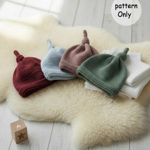 May include: Four knitted baby hats in burgundy, light blue, mauve, and green, each with a knotted top. The hats are displayed on a fluffy, cream-coloured surface, with a wooden block and the text "pattern Only".