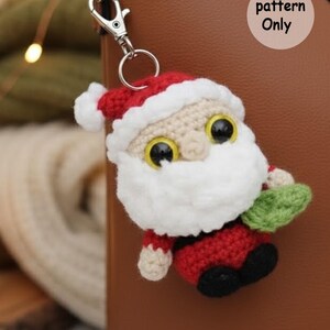 May include: Crocheted Santa Claus keychain with a red suit, white beard, and a red hat with white trim. The keychain has a silver clasp and a small green object in his hand. The text "pattern Only" is visible.