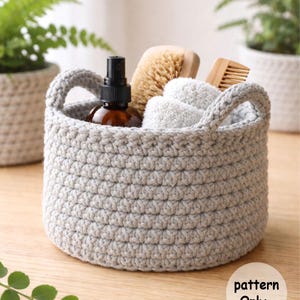 Puede incluir: Una cesta redonda de ganchillo gris claro con dos asas, llena de una botella marrón, un cepillo de madera, un peine y toallitas blancas. El texto "pattern Only" es visible en la superficie de madera.