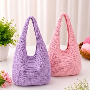 May include: Two handmade crochet handbags, one in a soft lavender purple and the other in a light pastel pink. Both bags feature a simple hobo-style silhouette with a single integrated handle and a consistent horizontal ribbed texture throughout.