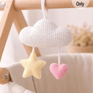 May include: A handmade crochet baby mobile featuring a white cloud, a yellow star, and a pink heart. The mobile is suspended from a wooden frame. The text "Pattern Only" is visible on the image.