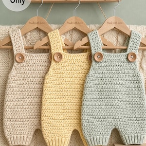 Puede incluir: Tres peleles de bebé de ganchillo hechos a mano en crema, amarillo y verde menta, cada uno con cierres de botones de madera. Los peleles cuelgan de perchas de madera que dicen "Made with Love". La imagen incluye el texto "Pattern Only".
