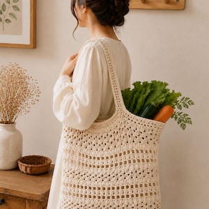 May include: A cream-colored crochet tote bag featuring an openwork pattern with alternating rows of solid stitches and square cutouts. The bag has two shoulder straps and is filled with fresh green lettuce and a single orange carrot.