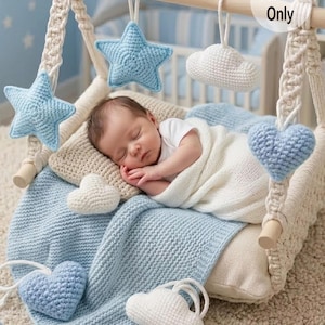 May include: A baby sleeps peacefully in a crocheted swing, surrounded by soft, handmade decorations. The swing features blue star and heart ornaments, along with white cloud accents. A blue knitted blanket and pillow provide comfort. The text "Pattern Only" is visible.