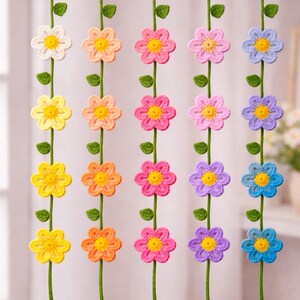 May include: Crocheted flower garland in various colors, including yellow, pink, and blue. Each flower has a yellow center and is strung on a green vine with green leaves. The garland hangs from a wooden rod.