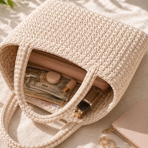 May include: Cream-colored, crocheted tote bag with two handles. The bag is open, revealing a pink wallet, cash, and various cosmetic items. A small, matching wallet and gold rings are also visible.